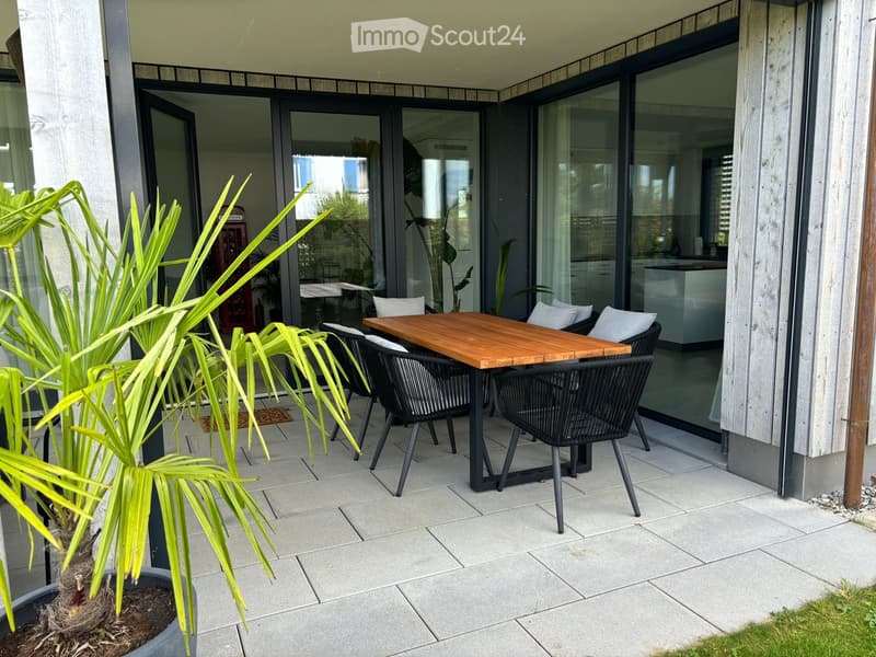 Balcony with a wooden table and black chairs, large sliding glass doors leading to the interior, and a potted plant on the left.