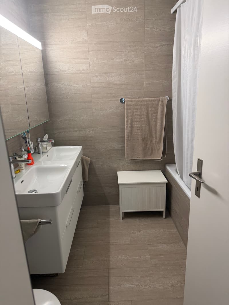 Modern bathroom with a double sink, mirror, towel, small stool, and bathtub. Light-colored tiles and wooden flooring.