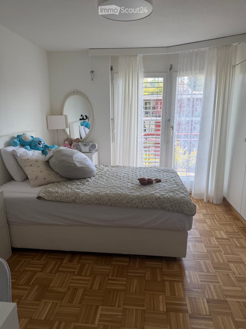 Cozy bedroom with wooden flooring, large bed, pillows, mirror, lamp, and toys.