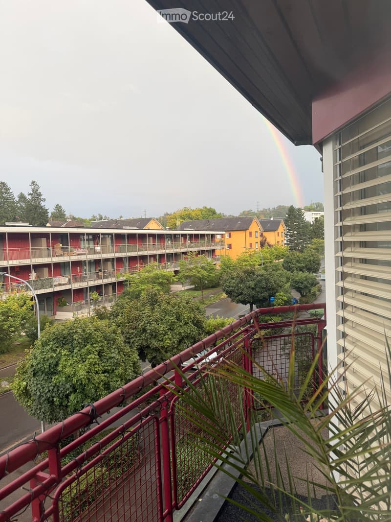 Balcony with red metal railing, overlooking a residential area with buildings, trees, and a visible rainbow in the sky.