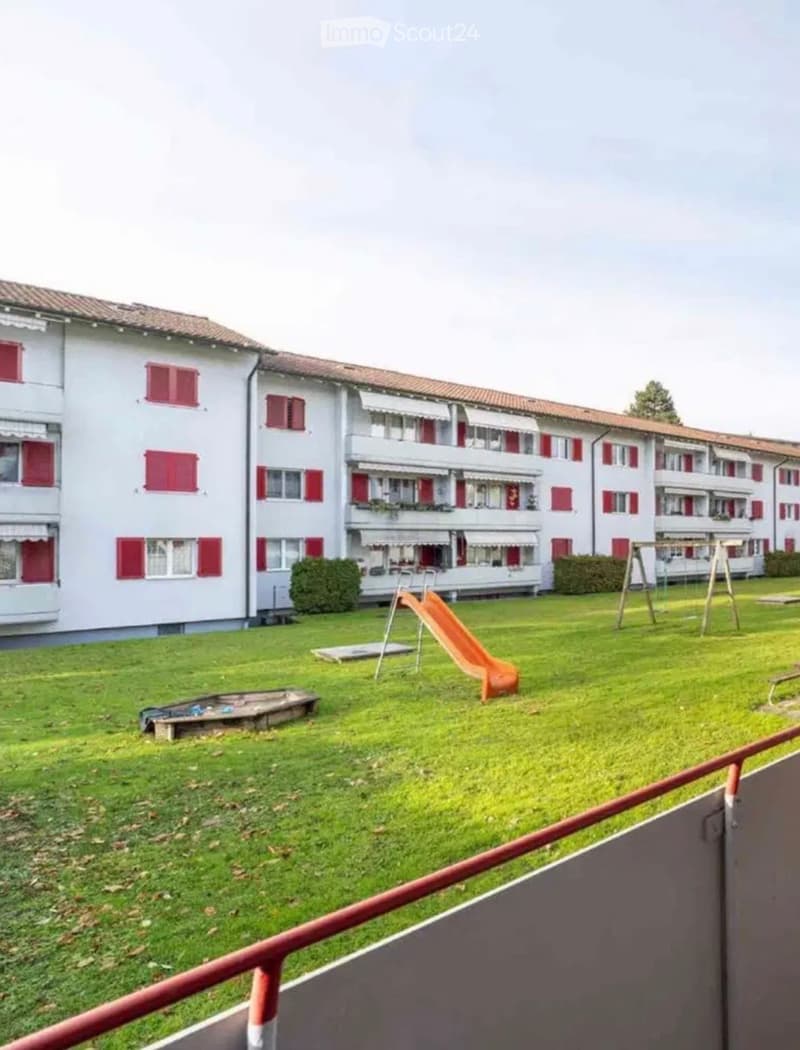 A grassy area in front of a multi-story residential building with red shutters, featuring a playground with a slide and swings.