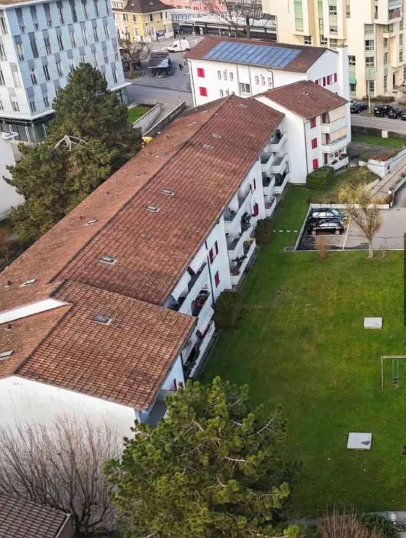 Aerial view of a multi-story residential building complex with red-tiled roofs, balconies, and surrounding greenery, including a lawn and parking area.