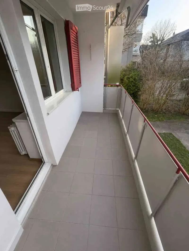 Balcony with tiled floor, white walls, red shutters, and a metal railing overlooking a garden area.