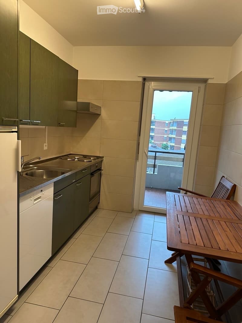 Modern kitchen with stainless steel appliances, a small wooden table and chairs, and a sliding door leading to a balcony.
