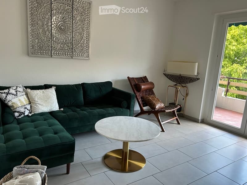 Modern living room with a green velvet sectional sofa, white coffee table with gold base, wooden chair, floor lamp, and decorative wall art. Large window with balcony access.