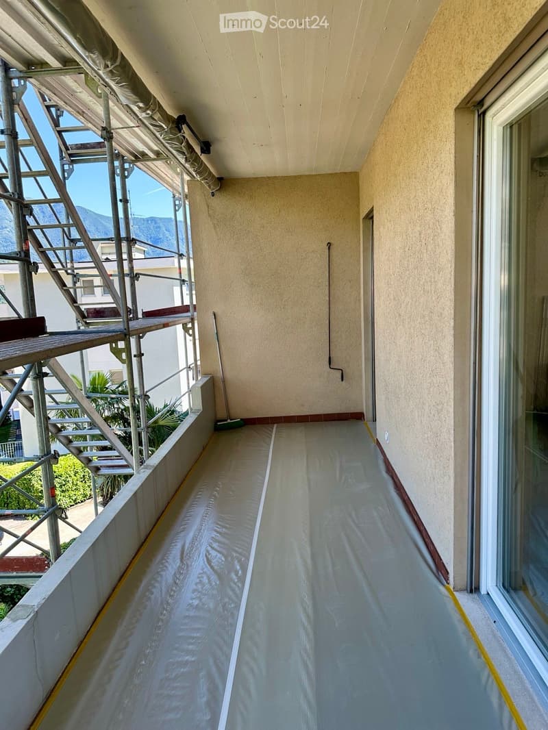 Balcony with a smooth, light-colored floor, beige walls, and a metal staircase on the left side. The balcony is partially covered with protective plastic sheeting and has a view of greenery and mountains in the background.