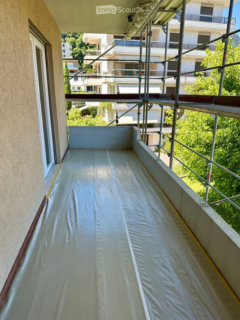 Balcony under construction with scaffolding and protective covering, overlooking a residential area with greenery and neighboring buildings.