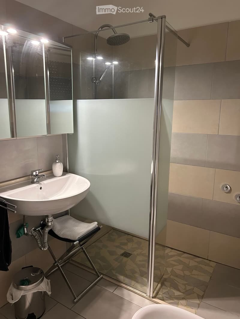 Modern bathroom with a white sink, mirror, glass-enclosed shower, and a small stool holding a folded towel. The walls are tiled in beige and grey, and the floor has a geometric tile pattern.