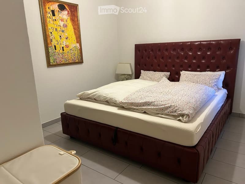 Cozy bedroom with a large upholstered bed, patterned bedding, a nightstand with a lamp, and a framed painting on the wall.