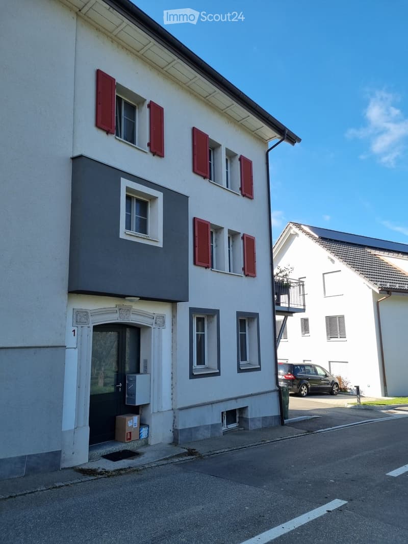 Two-story residential building with white walls, red shutters, and a gray accent wall on the left side. The entrance features a decorative archway and a small utility box. A car is parked nearby, and a neighboring building is visible in the background.