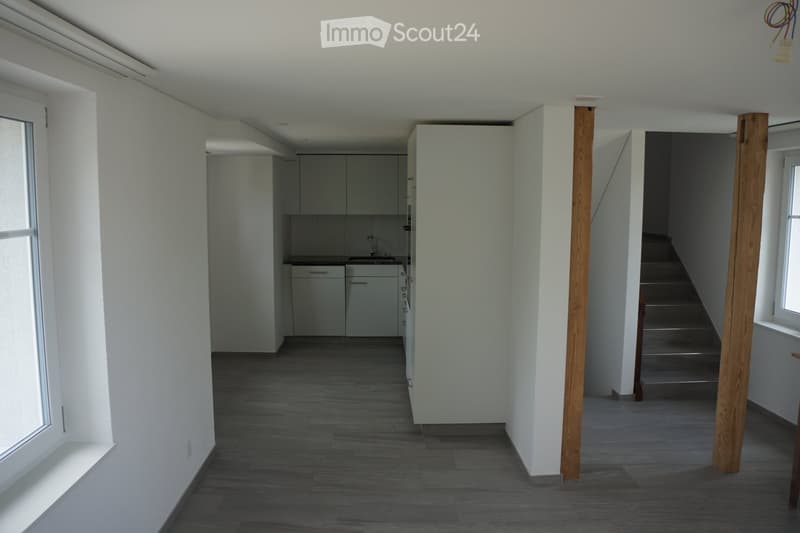 Empty hallway with wooden flooring, white walls, and a staircase leading upwards. A small kitchen area is visible to the left with cabinets and a sink.