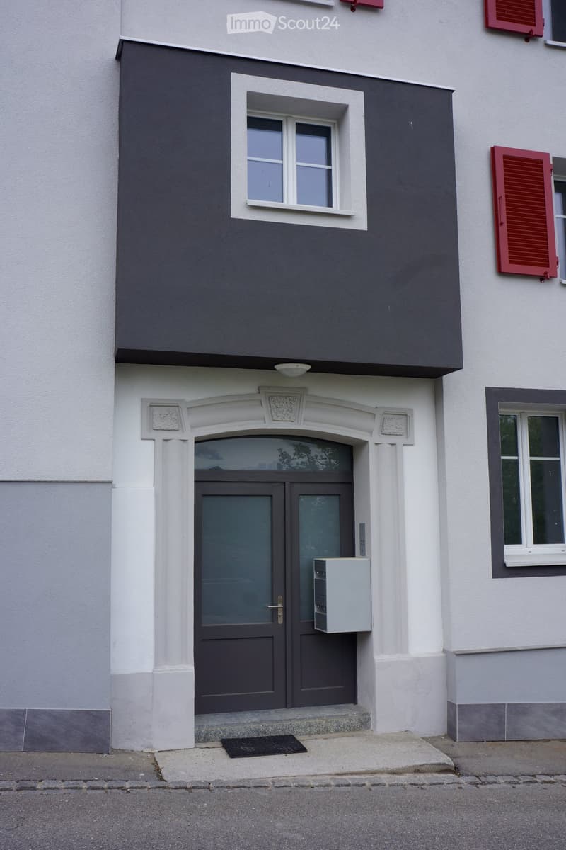 Two-story building with white and grey facade, double glass doors, and windows with shutters.
