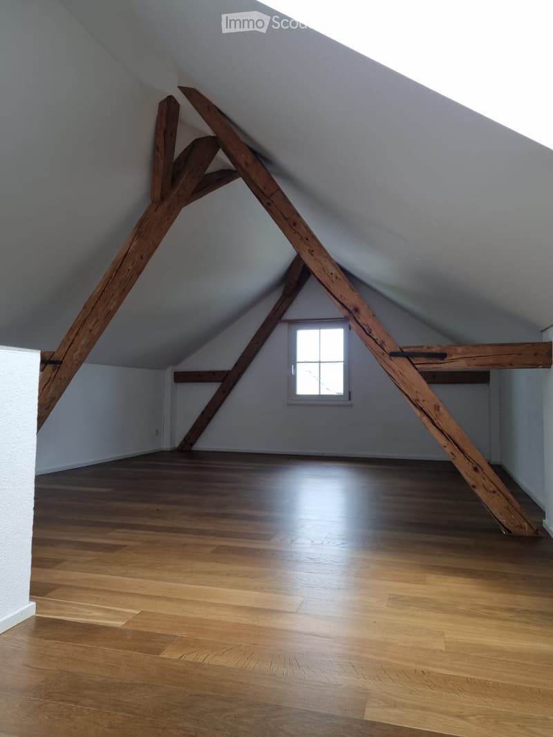 An attic space with wooden beams, wooden flooring, and a small window letting in natural light. The room is completely empty with no furniture or equipment.