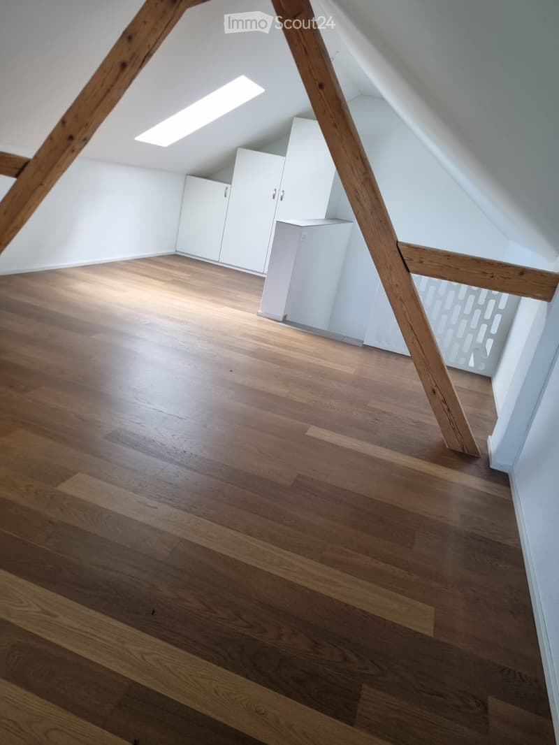 An empty attic room with wooden flooring, white walls, and built-in white cabinets. The room features exposed wooden beams and a skylight window allowing natural light to enter.
