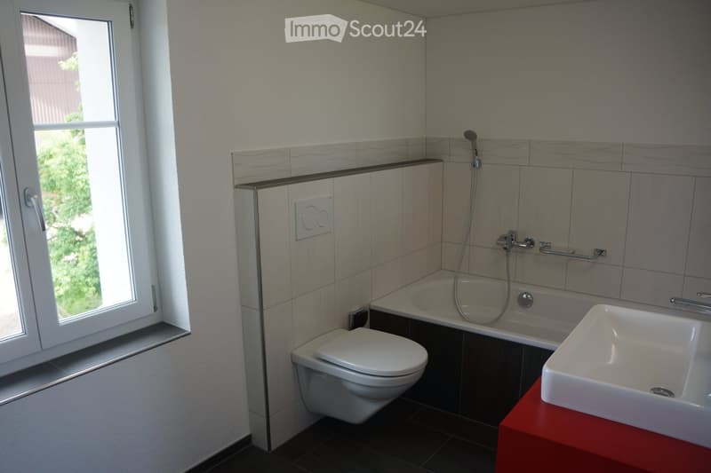 Modern bathroom with a toilet, bathtub, sink, and showerhead. The room features a large window with a view of greenery outside, and the walls are tiled with a light, textured pattern.