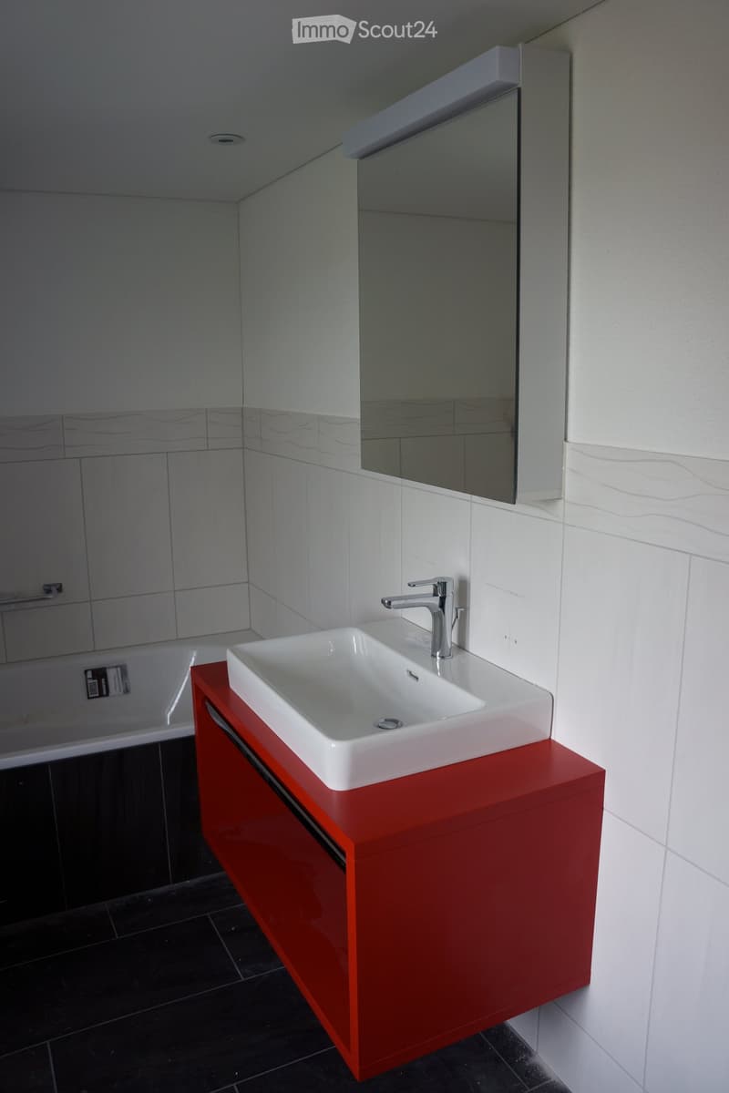 Modern bathroom with a white sink on a red cabinet, chrome faucet, large mirror, and white tiled walls.