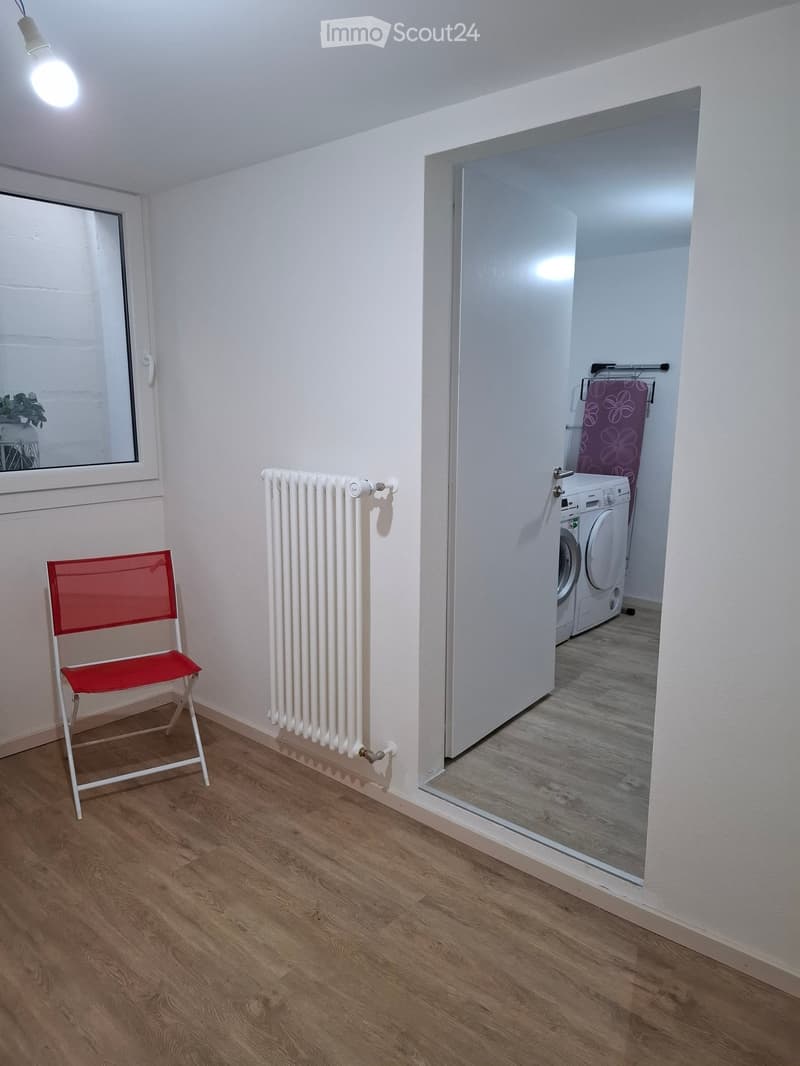 A small, well-lit room with wooden flooring, a white radiator, a red folding chair, and an open doorway leading to a laundry area with a washing machine and dryer.
