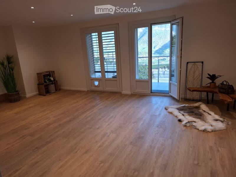 Bright, open room with wooden flooring, large windows, and a balcony view. Includes a bench, a small table with a handbag, a furry rug, a shelf with plants, and a decorative metal wall piece.