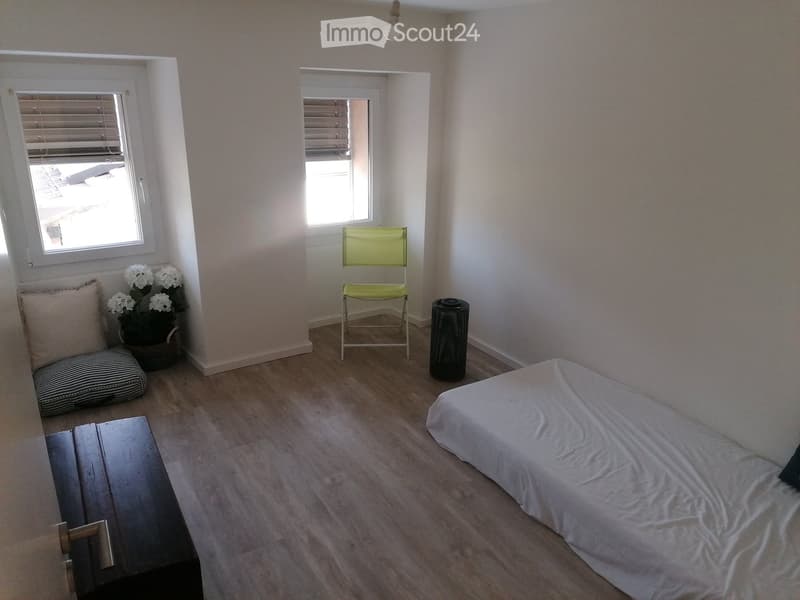 A small, well-lit bedroom with a single bed, a wooden chair, a small table, and a flower pot on a windowsill. The room features light-colored walls and wooden flooring.