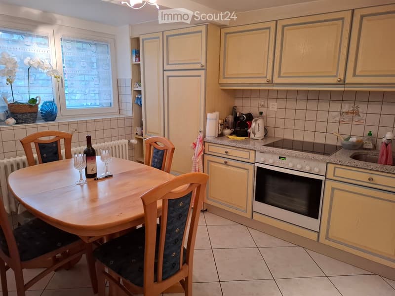 Bright kitchen with light wooden cabinets, a round wooden dining table with four chairs, and various appliances including an oven, refrigerator, coffee maker, and dishwasher. The counter has a few items including a bottle, cups, and a coffee maker.