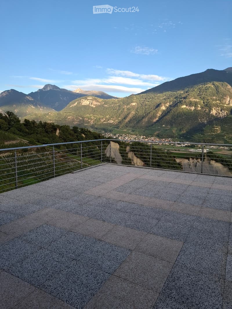 A spacious balcony with a modern metal railing, overlooking a scenic valley and mountains.