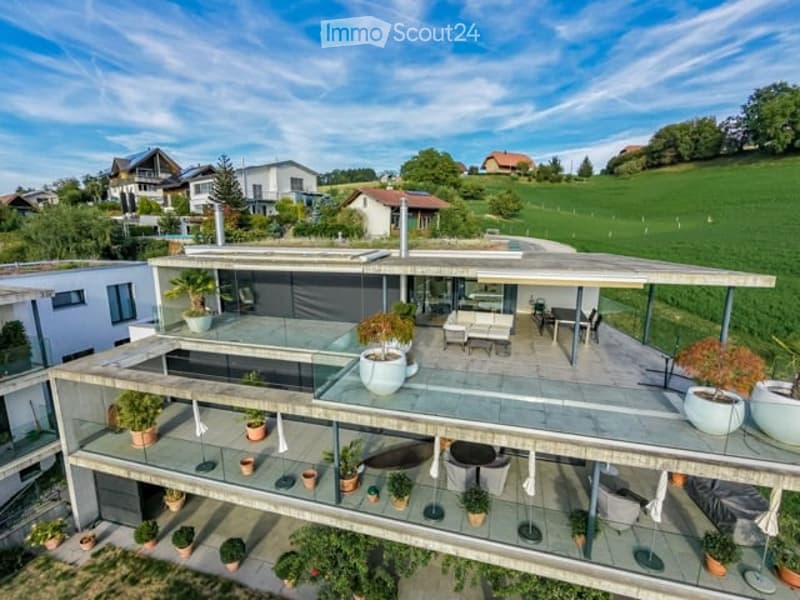 Modern two-story house with a large terrace and glass railings, surrounded by greenery and potted plants. The house features a flat roof and contemporary design.