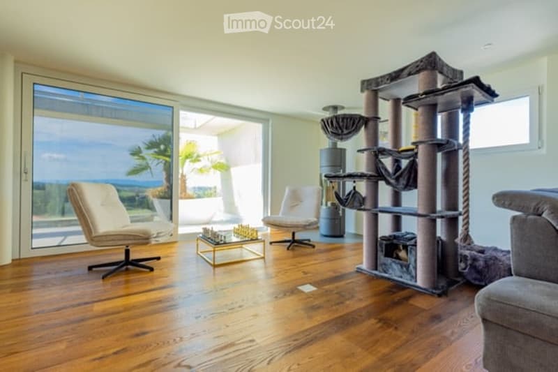 Bright living room with large windows offering a scenic ocean view, featuring a cream-colored sofa, two white armchairs, a multi-level cat tree, a small coffee table with a chessboard, and a polished wooden floor.