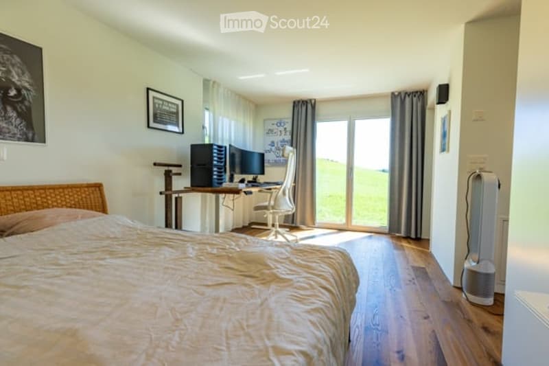 Well-lit bedroom with a large bed, a modern desk setup with a computer and dual monitors, a comfortable chair, and large windows with curtains. The room features light-colored wooden flooring and walls adorned with framed pictures.
