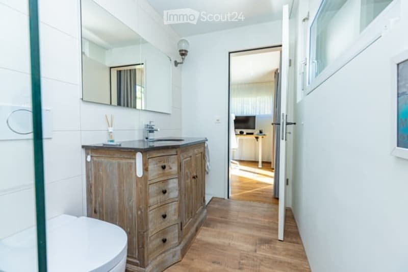 Bathroom with a wooden vanity, sink, mirror, and a visible toilet. The room has white tiled walls and a wooden floor.