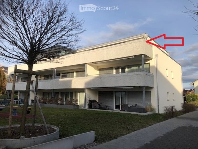 Modern two-story white building with balconies and large windows, featuring a raised ground floor and a small garden area.