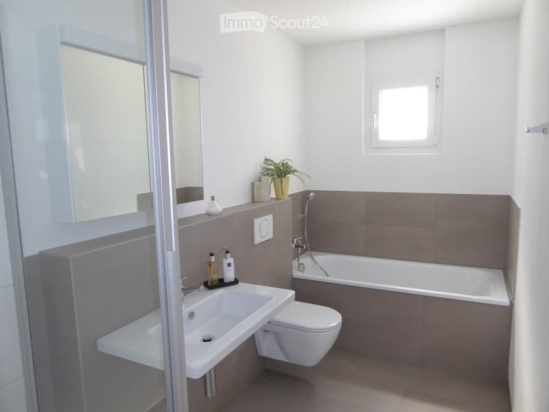 Modern bathroom with a white sink, toilet, bathtub, and a small plant on the windowsill. The walls are tiled in a neutral color scheme, and there is a large mirror above the sink.