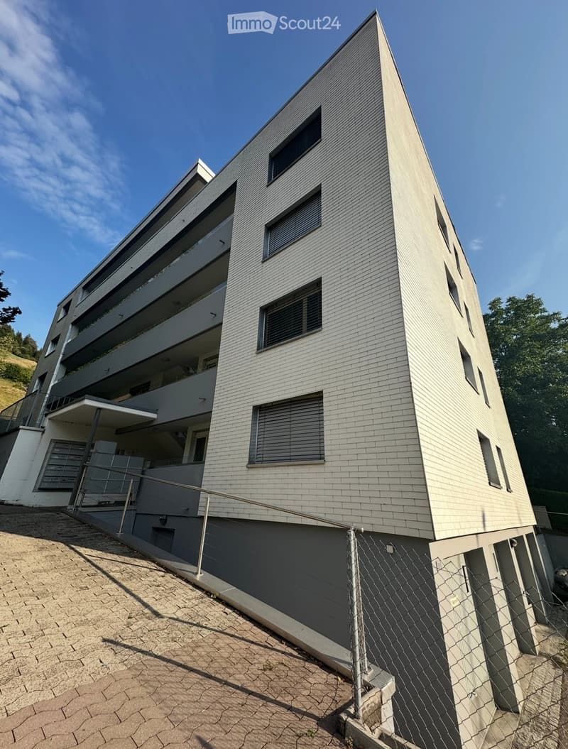 Modern multi-story building with grey brick facade, large windows, and balconies, situated on a raised ground floor with a paved entrance area.