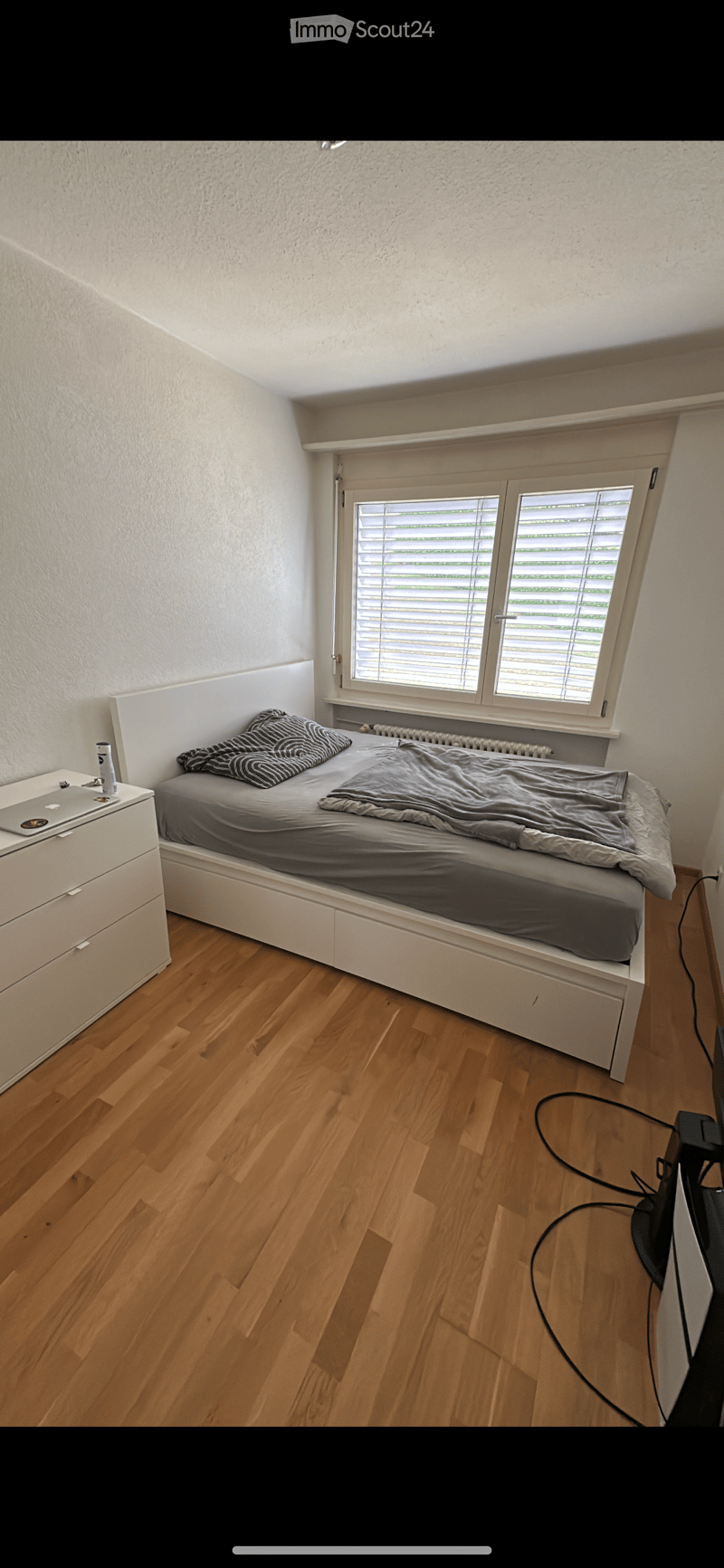 A modern, minimalist bedroom with a single bed, nightstand, laptop, and laptop charger. The room features wooden flooring and white walls.
