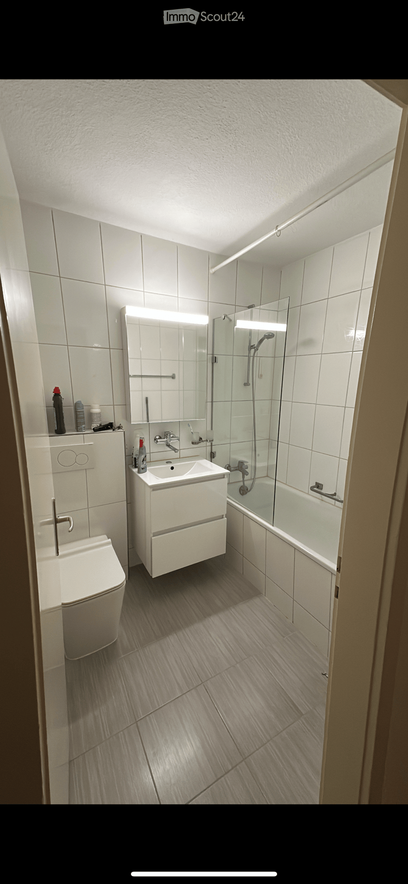 Modern bathroom with white tiles, a toilet, a sink with a mirror, a bathtub with a glass shower stall, and a light fixture above the mirror.
