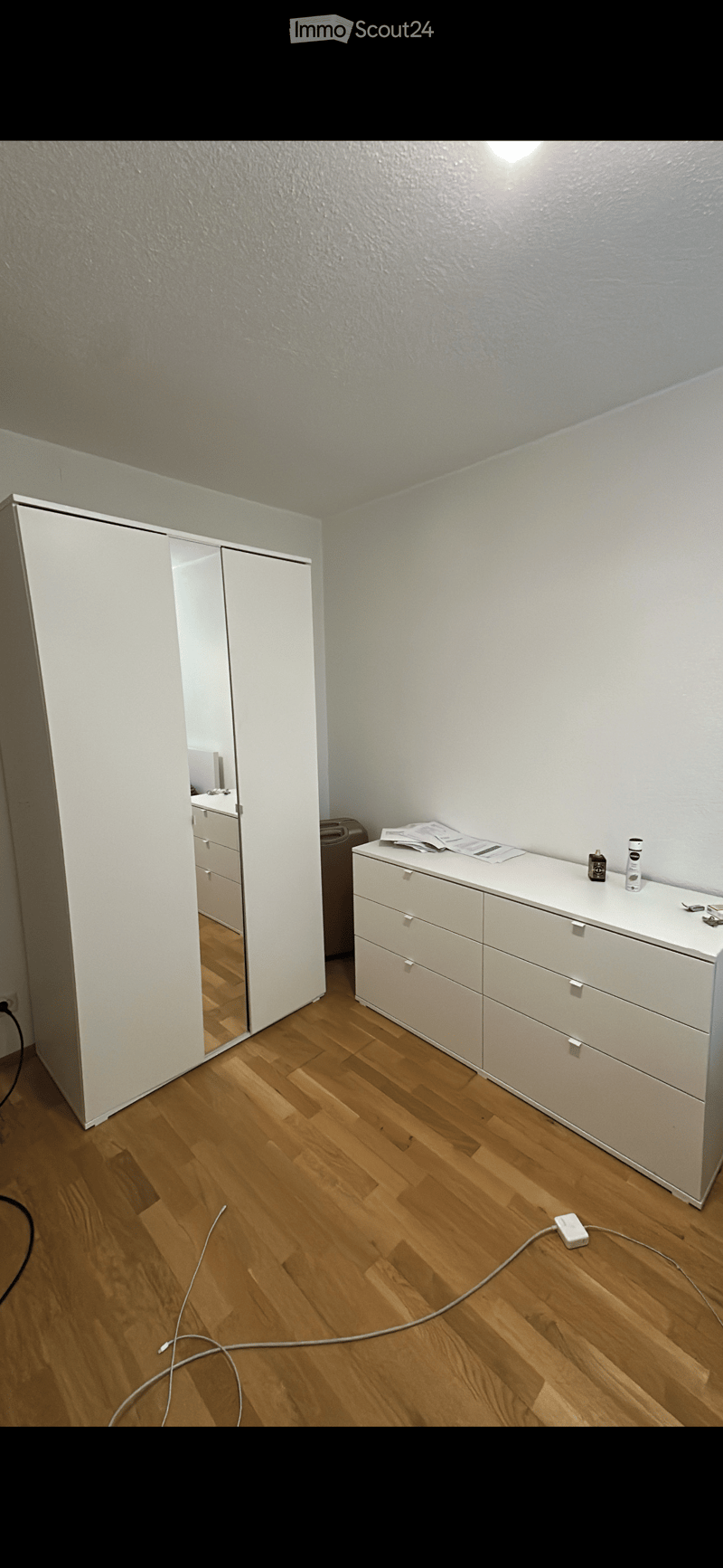A furnished room with a large white wardrobe, a white drawer unit, and a mirror. The room features wooden flooring and white walls.