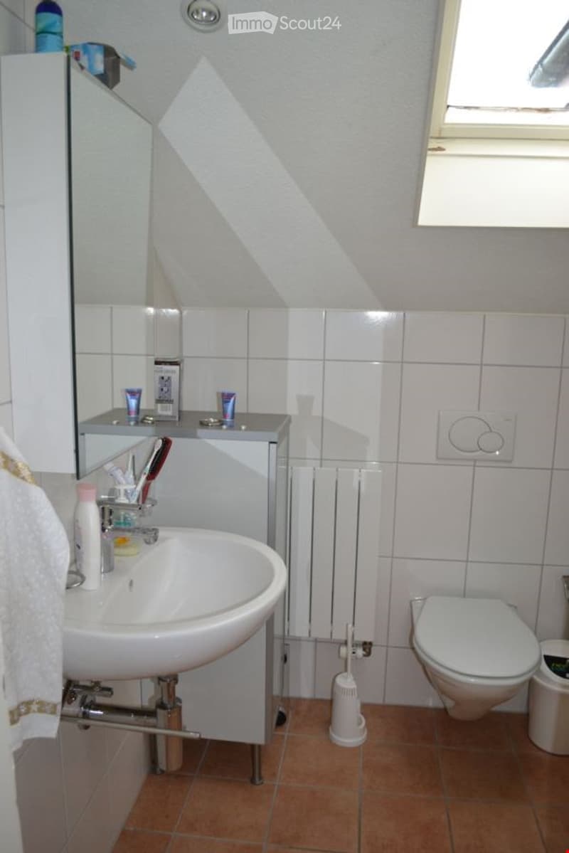 A small bathroom with a white sink, toilet, mirror, radiator, and various toiletries on the sink counter. The walls are tiled, and the floor is covered with brown tiles.
