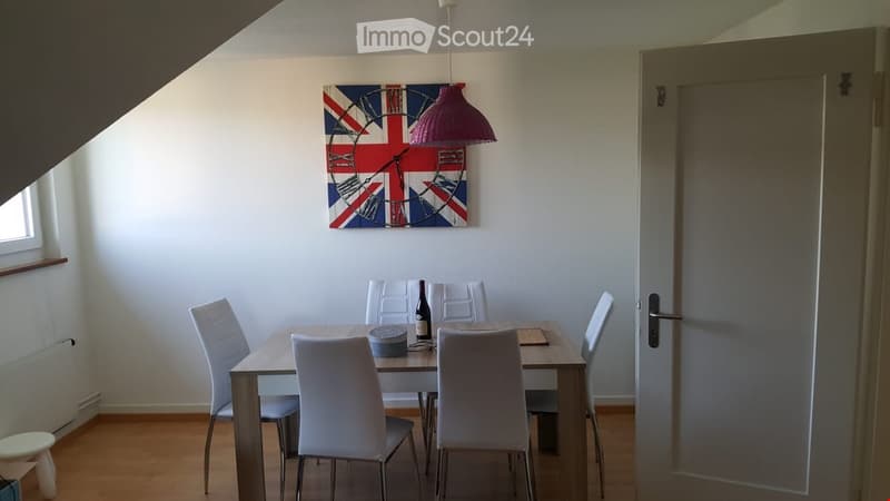 Dining room with a wooden table, six white chairs, a bottle on the table, a small stool, and a large wall clock with a Union Jack design.