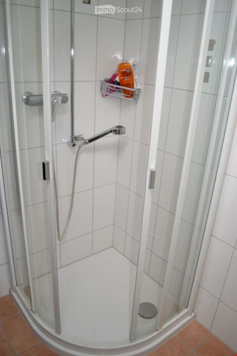 Modern walk-in shower with glass enclosure, tiled walls, and a built-in shelf holding a bottle of shower gel.