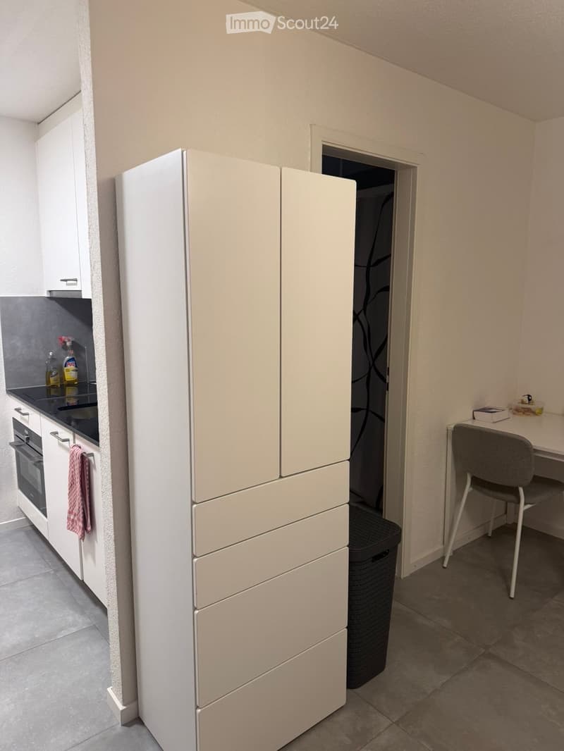 A small, modern kitchen with a refrigerator, oven, and a small desk area with a chair.