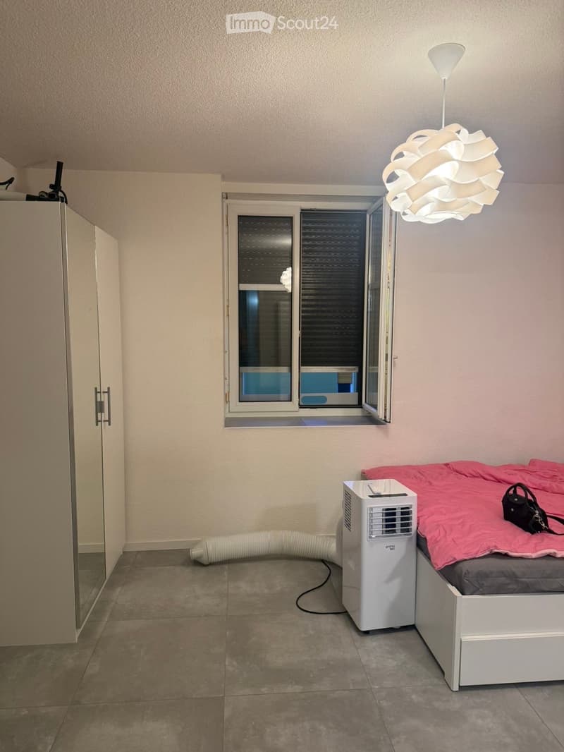 A small bedroom with a bed, a handbag on the bed, an air conditioner, and a modern lamp hanging from the ceiling.