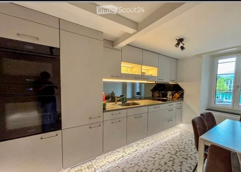 Modern kitchen with light grey cabinets, a built-in oven, a sink with a faucet, and a small dining area with a table and chairs.