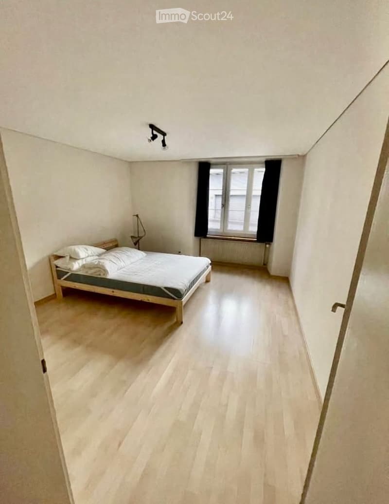 A small, unfurnished bedroom with a wooden bed, pillows, and a lamp near the window. The room has light wooden flooring and white walls.
