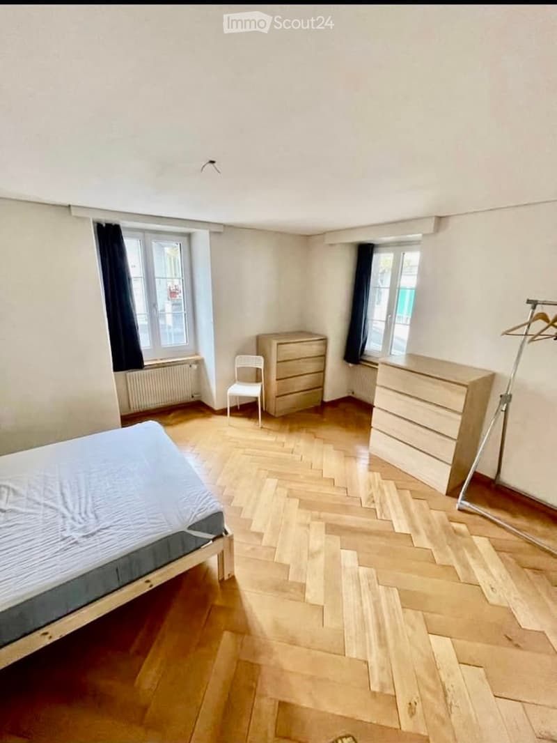 Bedroom with wooden herringbone flooring, a single bed, a white chair, a dresser, and a wardrobe. Two windows with dark curtains and radiators beneath them.