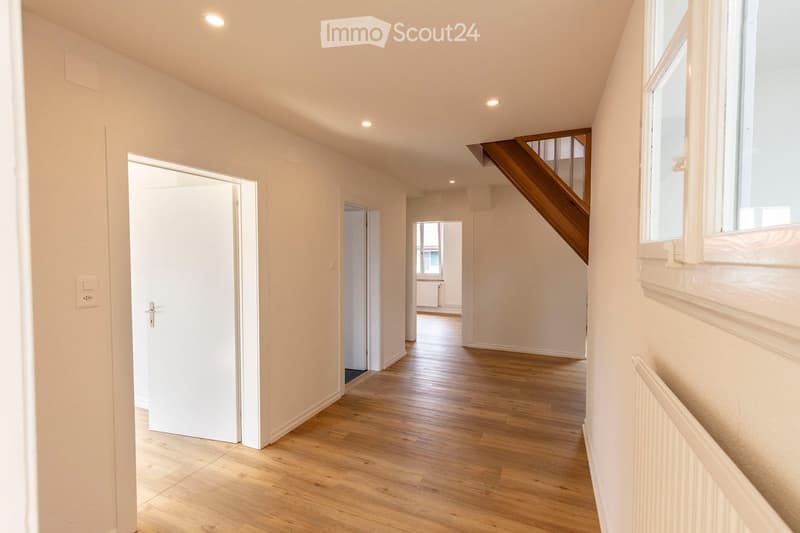 Bright hallway with wooden flooring, white walls, and recessed lighting. Features a staircase with wooden railing and several doors leading to other rooms.
