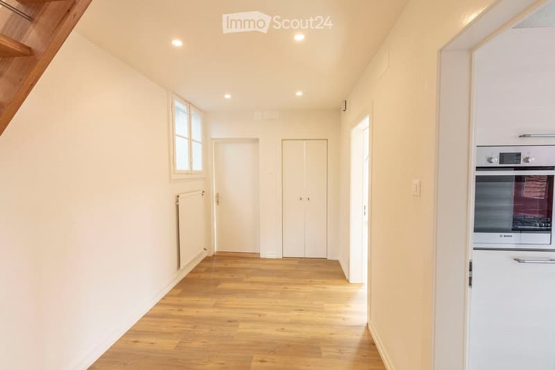 Bright hallway with wooden flooring, white walls, and recessed lighting. Features a window on the left, a radiator, a closed door, and a closet with double doors. An oven is visible in the adjacent kitchen area.