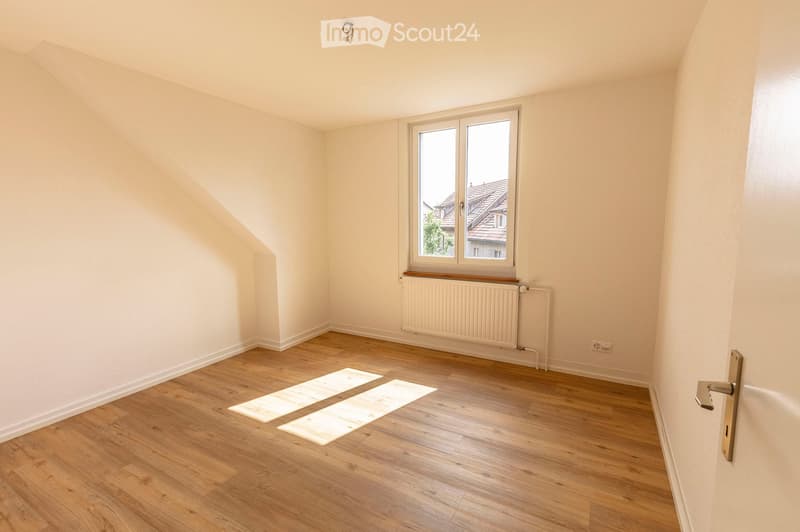 Empty room with wooden flooring, white walls, a window with a radiator below, and a door with a golden handle.