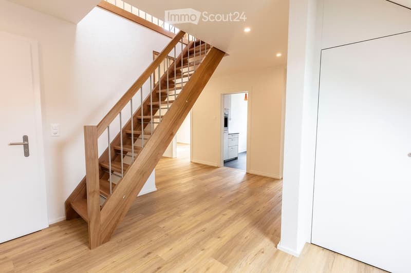 Spacious hallway with wooden flooring, a wooden staircase with metal railings, and a white door leading to another room.