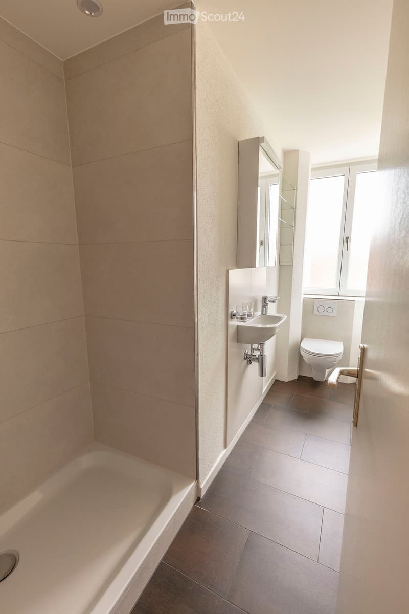 Modern bathroom with a bathtub, sink, toilet, and mirror. Light-colored tiles and a window provide natural light.