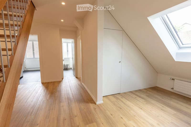 Bright, modern hallway with wooden flooring, white walls, and a staircase leading upwards. Features a built-in closet and a skylight window at the end.
