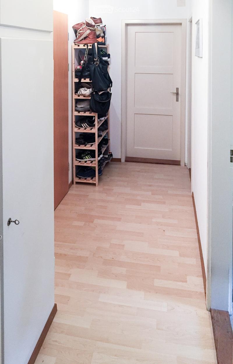 A narrow hallway with light wooden flooring and white walls. A wooden shoe rack with multiple pairs of shoes and bags is positioned against the left wall, and a closed white door is visible at the end of the hallway.